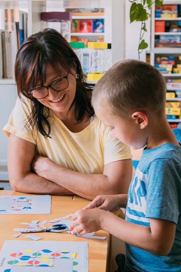 Einzeltherapie Angelika Reichartzeder und ein Bub sitzen bei einem Tisch: Der Bub schneidet und klebt Papierstücke zusammen.