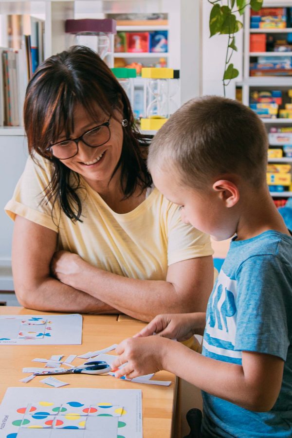 Angelika Reichartzeder und ein Bub sitzen bei einem Tisch: Der Bub schneidet und klebt Papierstücke zusammen.