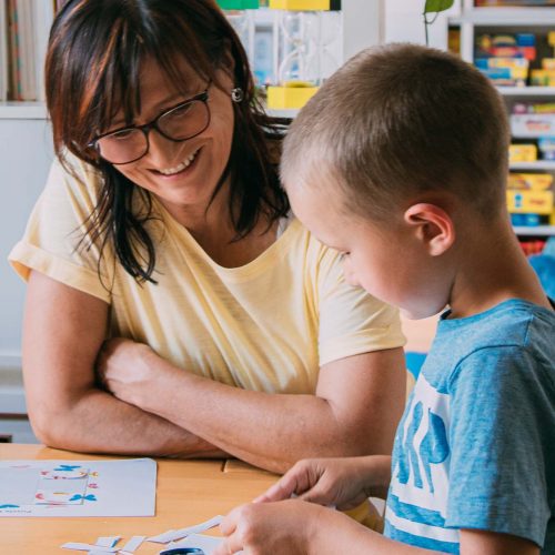 Einzeltherapie Angelika Reichartzeder und ein Bub sitzen bei einem Tisch: Der Bub schneidet und klebt Papierstücke zusammen.